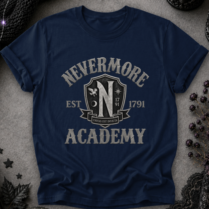 A navy blue short-sleeve t-shirt with a distressed off-white graphic reading 'NEVERMORE ACADEMY' and a crest-style emblem, laid flat on a textured gray surface with dark decorative props around it.