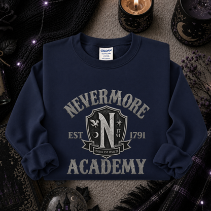 A navy blue crewneck sweatshirt with a distressed gray 'Nevermore Academy' graphic on the front, styled in a cozy, dark-themed flat-lay with candles, a knit blanket, and a black plaid scarf.