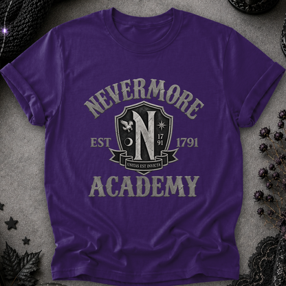 A purple short-sleeve t-shirt with a distressed gray graphic reading 'NEVERMORE ACADEMY' and a crest-style emblem, laid flat on a textured gray surface with dark decorative props around it.
