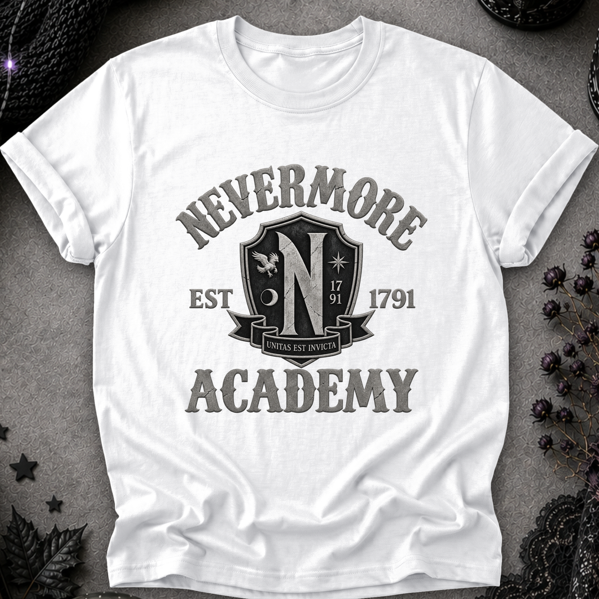 A white short-sleeve crewneck t-shirt with a distressed black graphic reading 'NEVERMORE ACADEMY' and a crest-style emblem, laid flat on a dark textured surface with decorative dried flowers and lace around it.