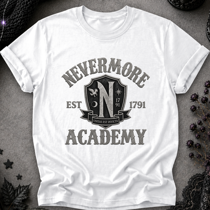 A white short-sleeve crewneck t-shirt with a distressed black graphic reading 'NEVERMORE ACADEMY' and a crest-style emblem, laid flat on a dark textured surface with decorative dried flowers and lace around it.