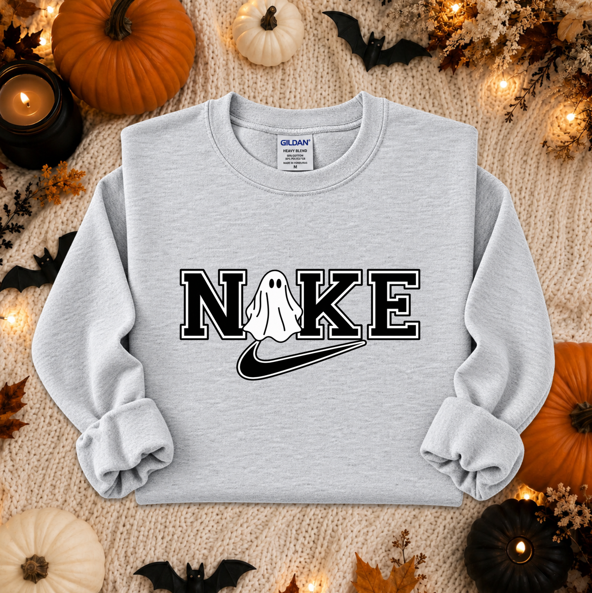 A light heather gray sweatshirt laid flat on a cozy beige knit blanket, surrounded by Halloween decorations including pumpkins, candles, leaves, and bats. The sweatshirt has a graphic print reading 'NIKE' with a ghost and a black swoosh, and a visible Gildan neck label.
