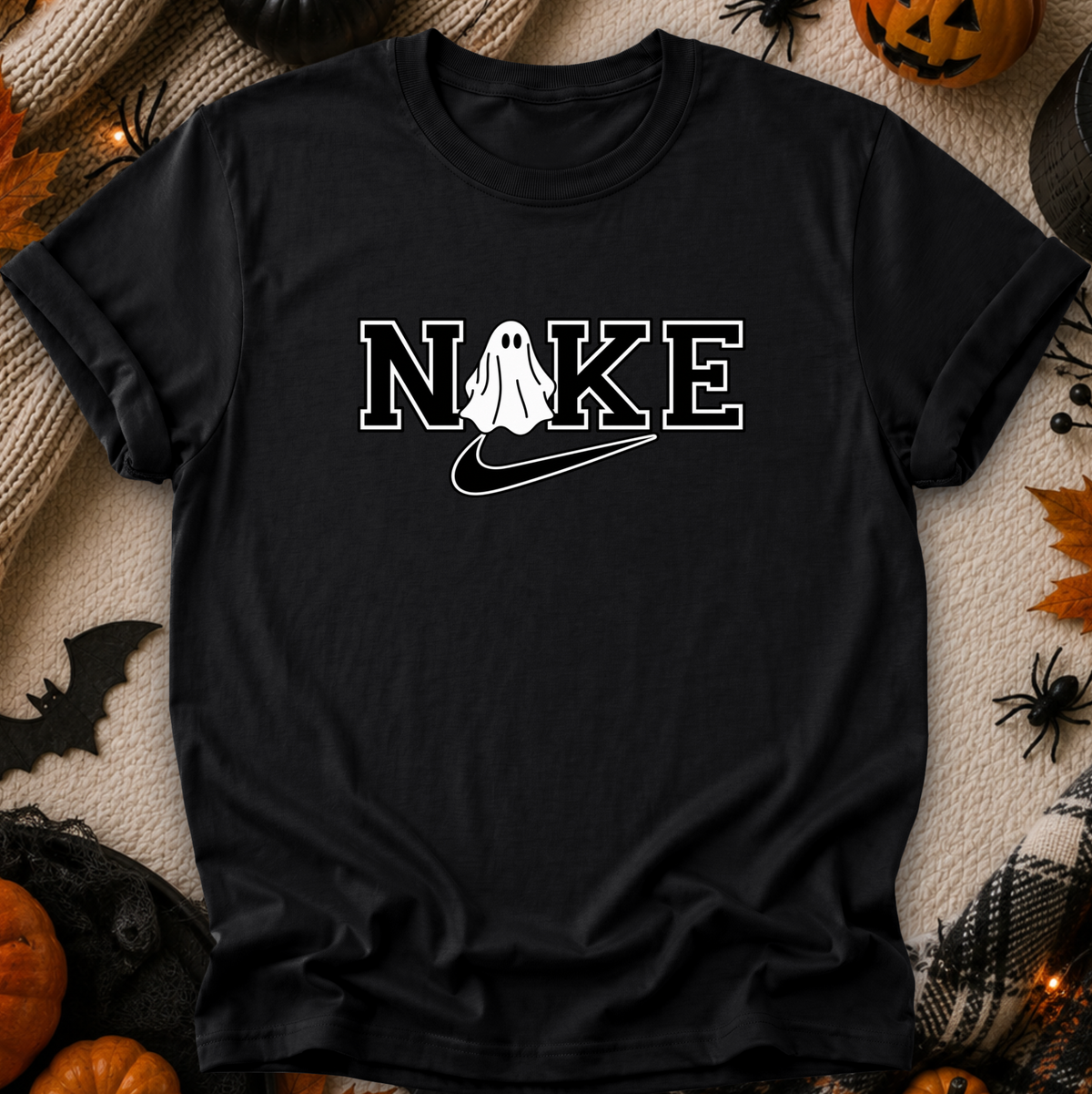 A black short-sleeve graphic T-shirt laid flat on a cozy beige knit blanket with Halloween decorations around it, including pumpkins, spiders, and a bat. The shirt has a white outlined 'NOKE' graphic across the chest with a ghost figure replacing the O and a white swoosh underneath.