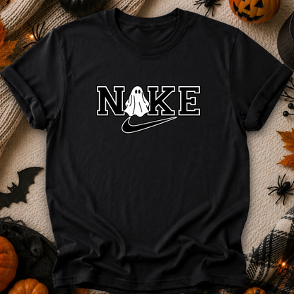 A black short-sleeve graphic T-shirt laid flat on a cozy beige knit blanket with Halloween decorations around it, including pumpkins, spiders, and a bat. The shirt has a white outlined 'NOKE' graphic across the chest with a ghost figure replacing the O and a white swoosh underneath.