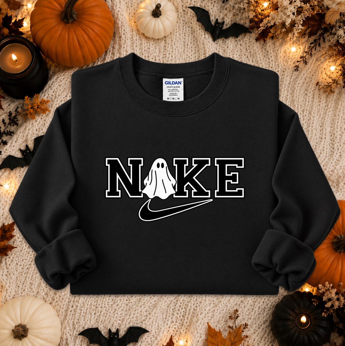 A black crewneck sweatshirt with a white graphic reading 'NAKE' where the O is replaced by a ghost and a swoosh, styled flat-lay on a beige knit blanket with Halloween props including pumpkins, bats, and candles.