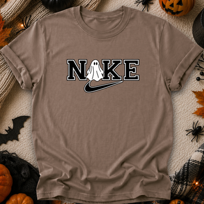 A taupe brown t-shirt with a 'N I K E' graphic featuring a white ghost and a black swoosh, styled in a flat-lay with Halloween props like pumpkins, a spider, and a witch hat.