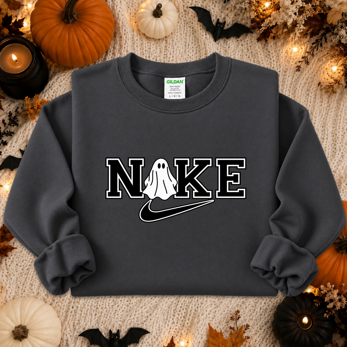 A dark charcoal gray sweatshirt with a 'NIKE' graphic featuring a ghost and a Nike swoosh, styled in a cozy autumn flat-lay with pumpkins, candles, and a knitted blanket background.