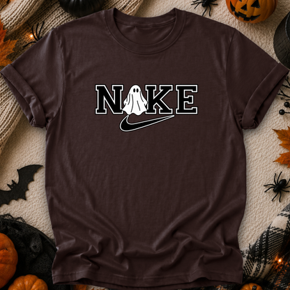 A black short-sleeve graphic T-shirt laid flat on a cozy beige knit blanket with Halloween decorations around it, including pumpkins, spiders, and a bat. The shirt has a white outlined 'NIKE' graphic across the chest with a ghost figure replacing the O and a white swoosh underneath.