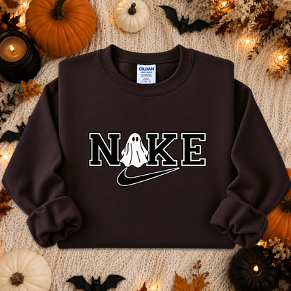 A dark brown crewneck sweatshirt with a white graphic reading 'NIKE' and a ghost-shaped 'I' over a swoosh, styled in a flat lay with autumn props like pumpkins, candles, and a knit blanket. A Gildan brand tag is visible inside the collar.
