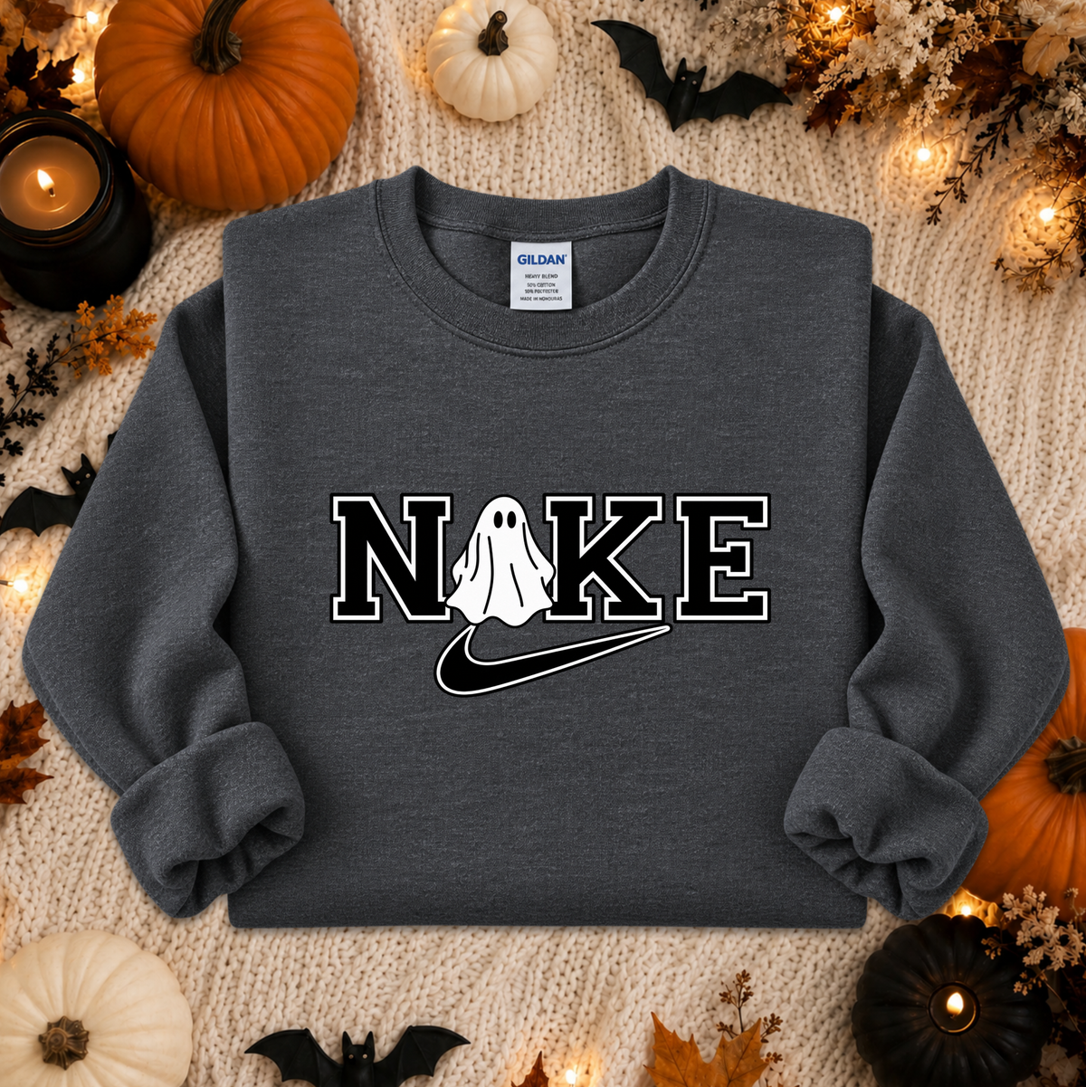A dark gray crewneck sweatshirt with a 'NIKE' graphic featuring a ghost and a swoosh, styled in a flat lay with Halloween decorations like pumpkins, bats, and candles.