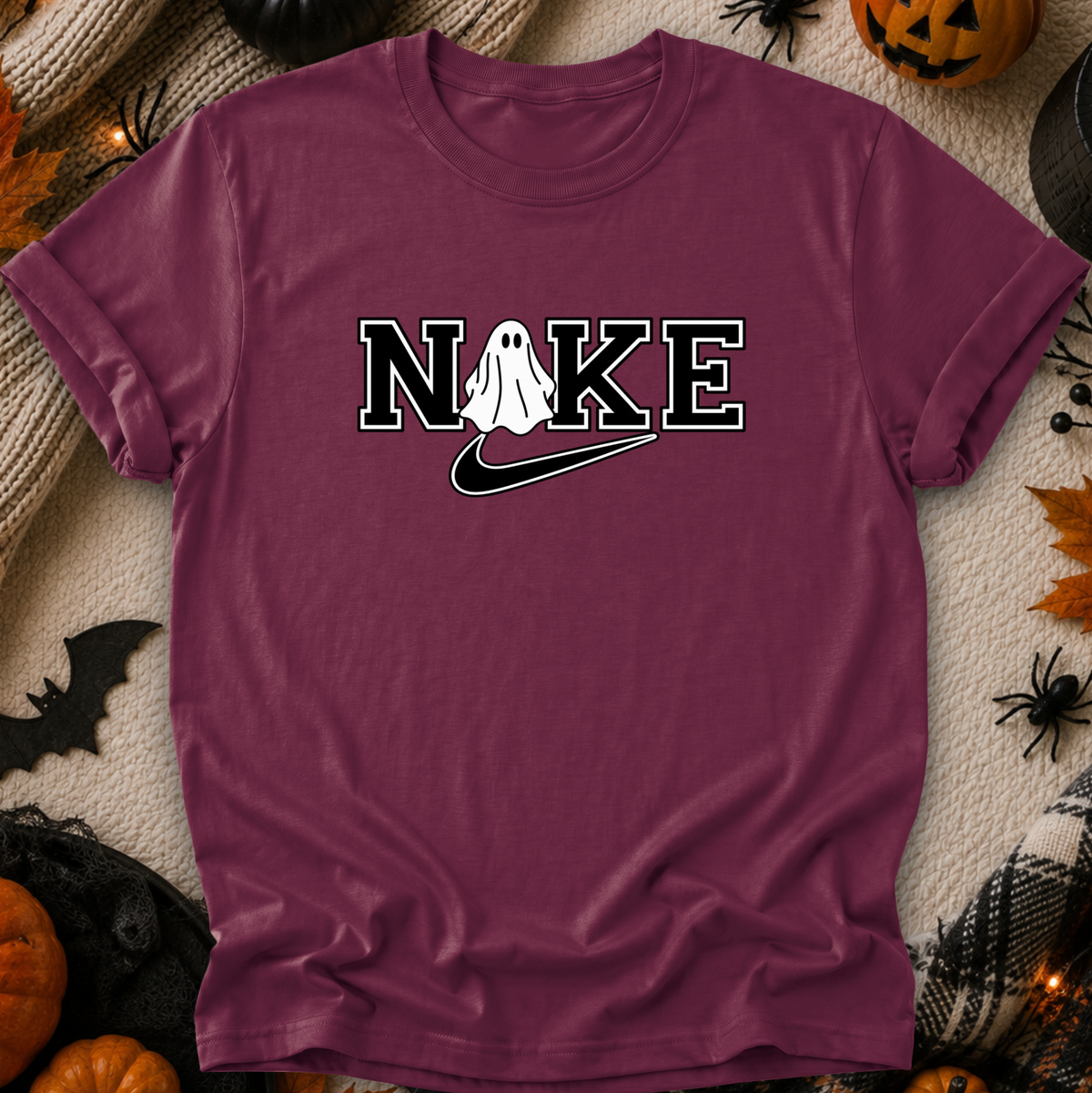 A maroon short-sleeve t-shirt with a 'N I K E' graphic featuring a white ghost and a black swoosh, styled in a flat-lay with Halloween decorations.