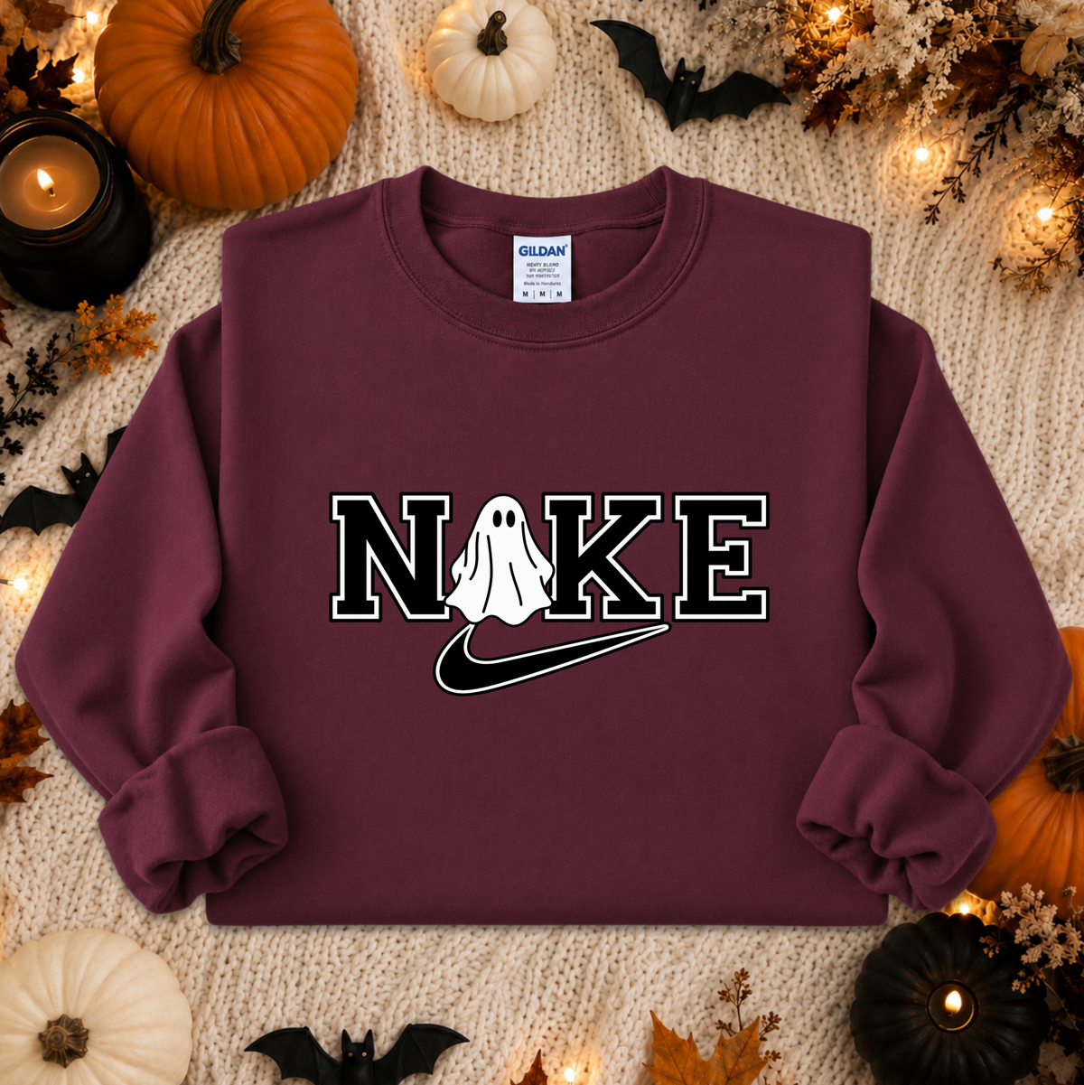 A maroon sweatshirt laid flat on a cream knit surface with Halloween props including pumpkins, bats, leaves, and candles. The sweatshirt has a large chest graphic reading 'NIKE' with a ghost over the O and a black Nike swoosh underneath, plus a visible Gildan neck label.