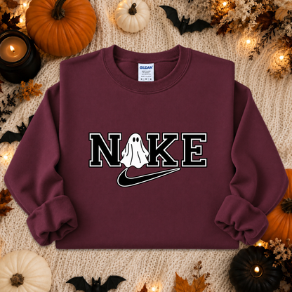 A maroon sweatshirt laid flat on a cream knit surface with Halloween props including pumpkins, bats, leaves, and candles. The sweatshirt has a large chest graphic reading 'NIKE' with a ghost over the O and a black Nike swoosh underneath, plus a visible Gildan neck label.