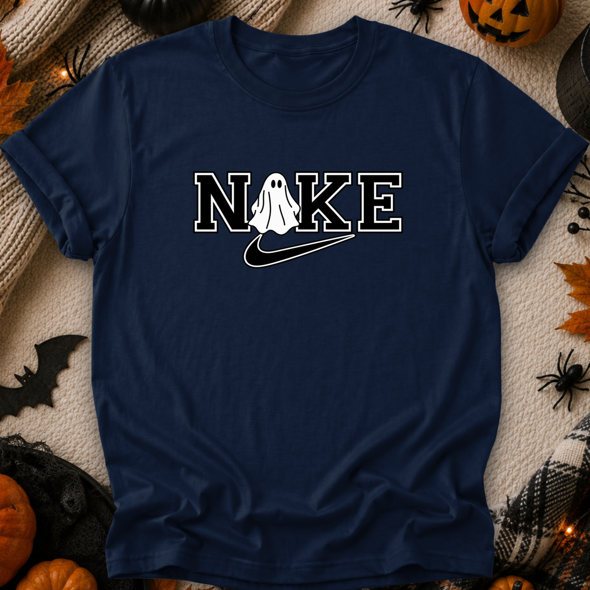 A navy blue t-shirt with a white graphic reading 'NIKE' where the 'O' is replaced by a ghost and a swoosh, styled in a flat lay with Halloween decorations like pumpkins, a spider, and a black cat toy on a beige blanket.