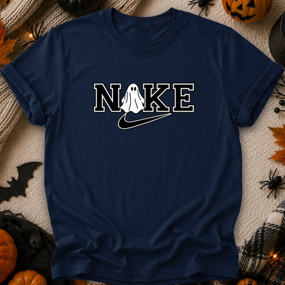 A navy blue t-shirt with a white graphic reading 'NIKE' where the 'O' is replaced by a ghost and a swoosh, styled in a flat lay with Halloween decorations like pumpkins, a spider, and a black cat toy on a beige blanket.