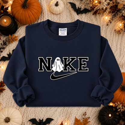 A navy blue crewneck sweatshirt with a graphic print reading 'NIKE' where the 'I' is replaced by a white ghost and a black swoosh. The sweatshirt is laid flat on a beige knit blanket surrounded by Halloween decorations including pumpkins, bats, and candles.