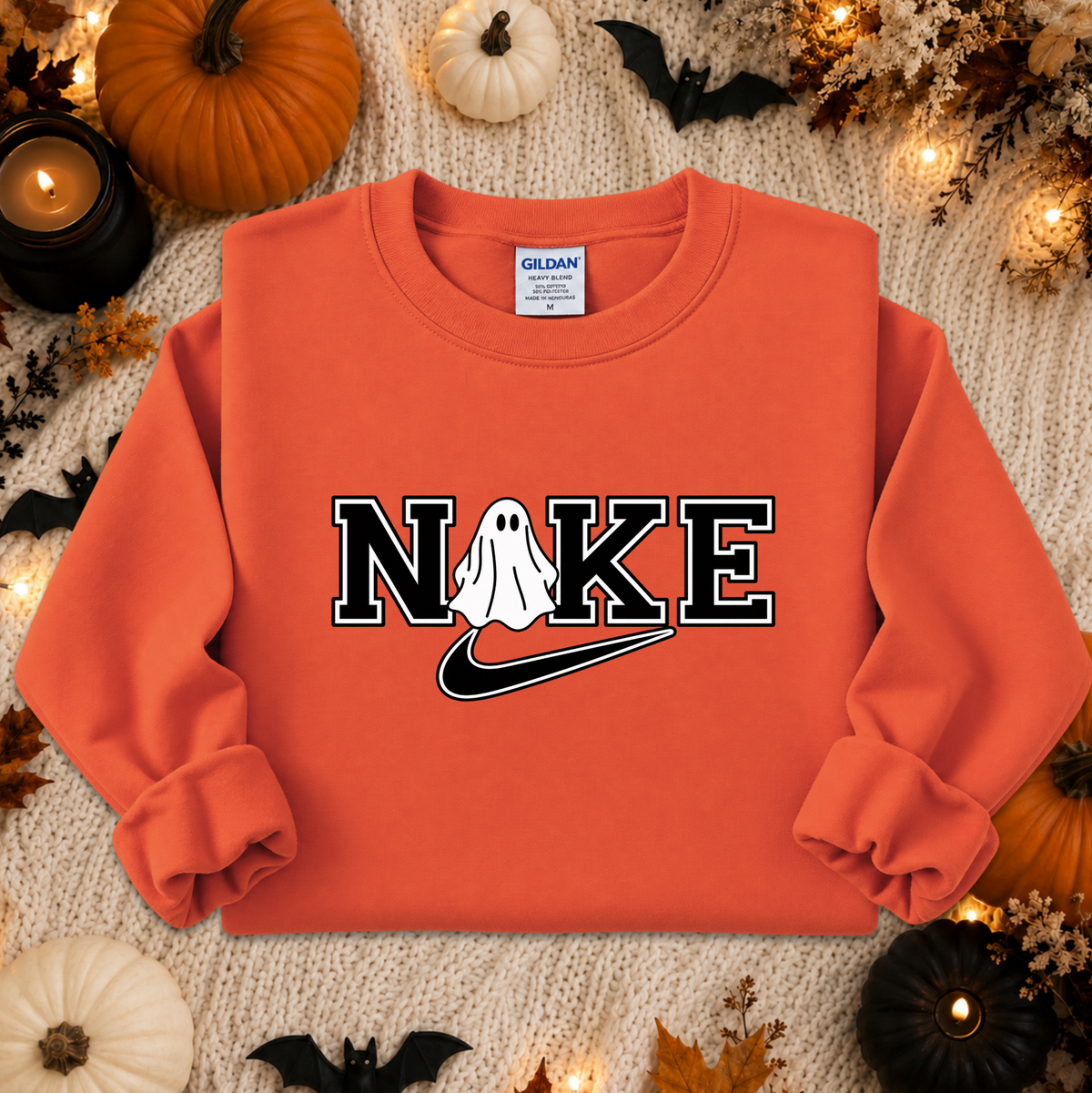 An orange crewneck sweatshirt with a 'NIKE' graphic featuring a ghost and a swoosh, styled in a flat lay with Halloween decorations like pumpkins, candles, and bats on a cream knit blanket.