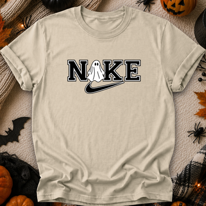 A beige graphic t-shirt laid flat on a cozy, Halloween-themed background. The shirt features a 'NIKE' parody graphic with a white ghost in place of the 'I', and a black swoosh underneath.