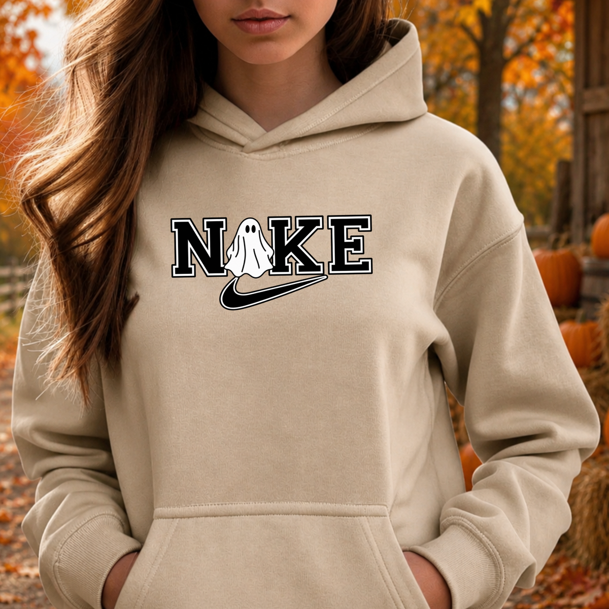 A woman wearing a beige hoodie with a graphic that reads 'N O K E' with a ghost and a swoosh, photographed outdoors in an autumn setting with pumpkins and trees in the background.