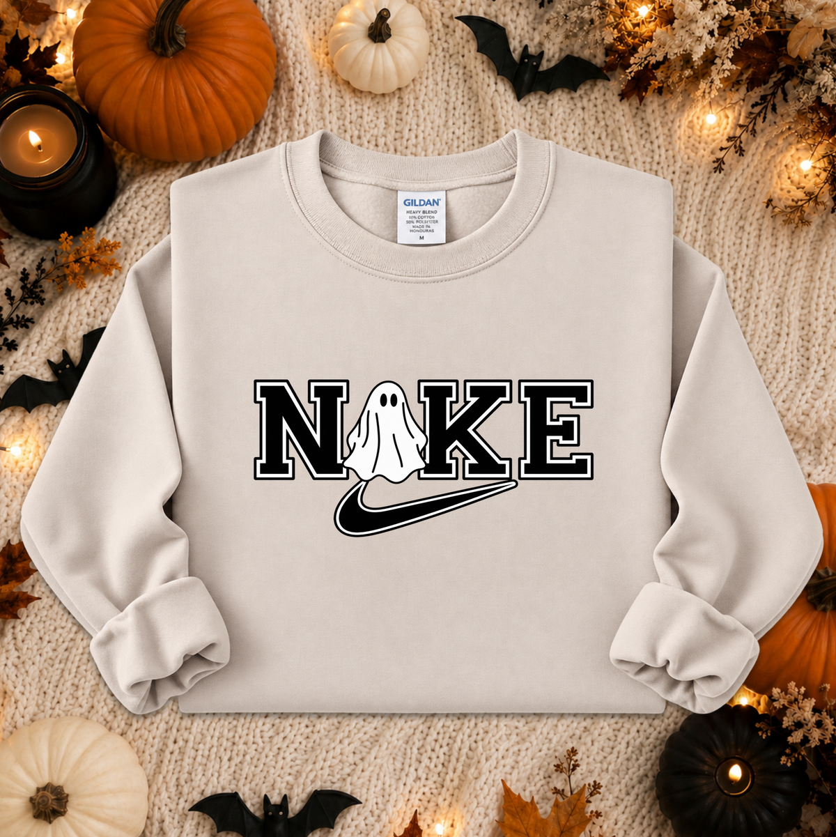A light beige crewneck sweatshirt with a 'NIKE' graphic featuring a ghost and a swoosh, styled in a flat lay with Halloween decorations like pumpkins, bats, and candles.