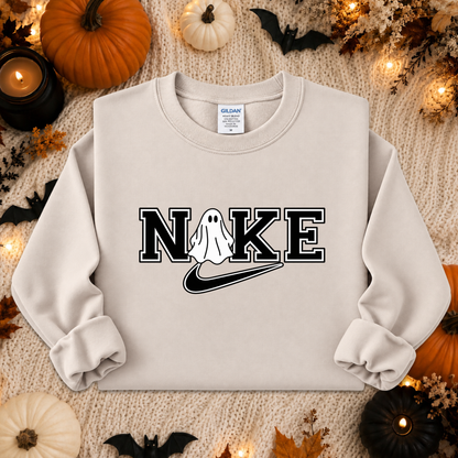 A light beige crewneck sweatshirt with a 'NIKE' graphic featuring a ghost and a swoosh, styled in a flat lay with Halloween decorations like pumpkins, bats, and candles.