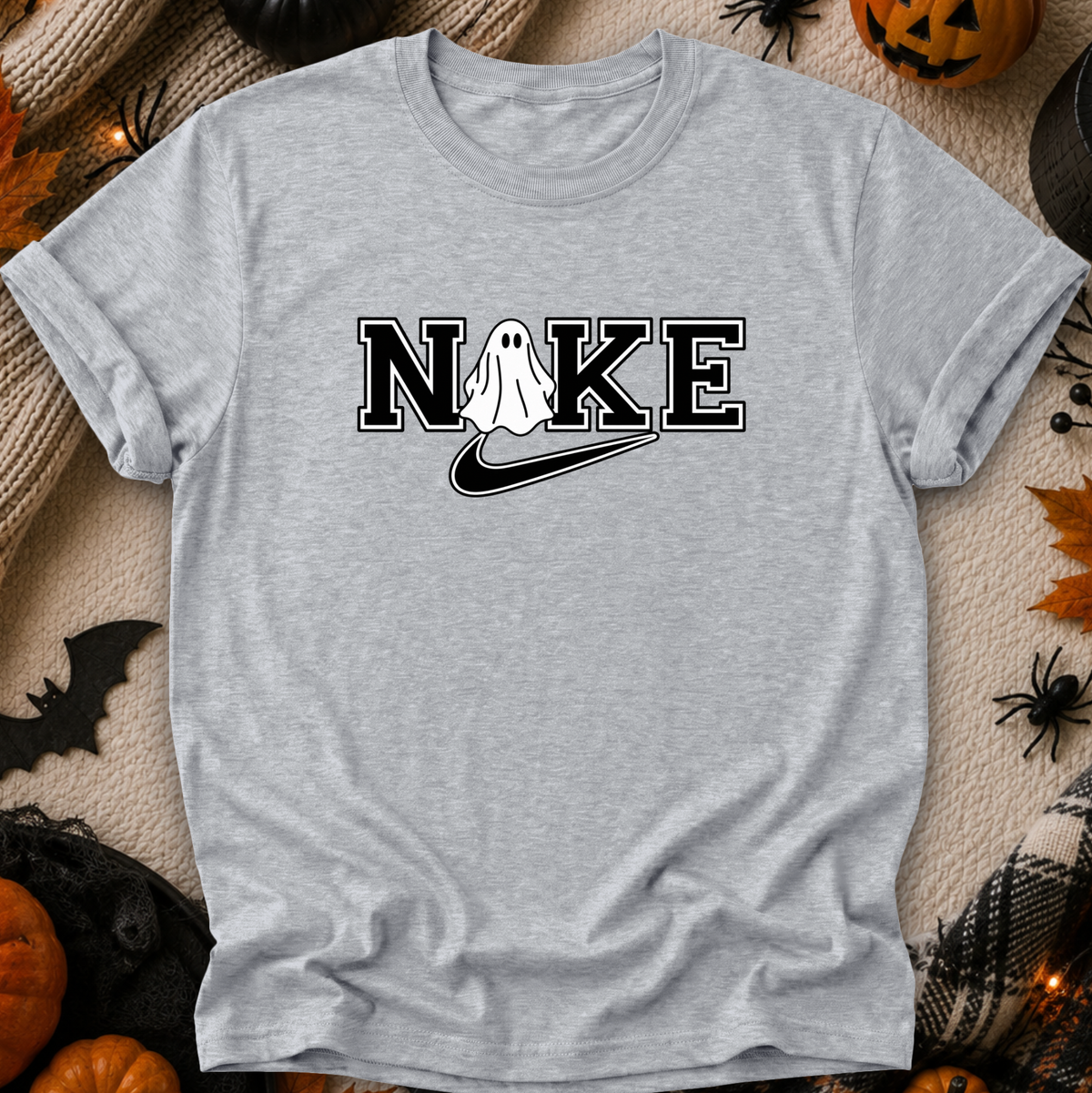 A light heather gray t-shirt with a 'N I K E' graphic featuring a ghost and a swoosh, laid flat on a cozy beige knit blanket surrounded by Halloween decorations like pumpkins, a spider, and a bat.