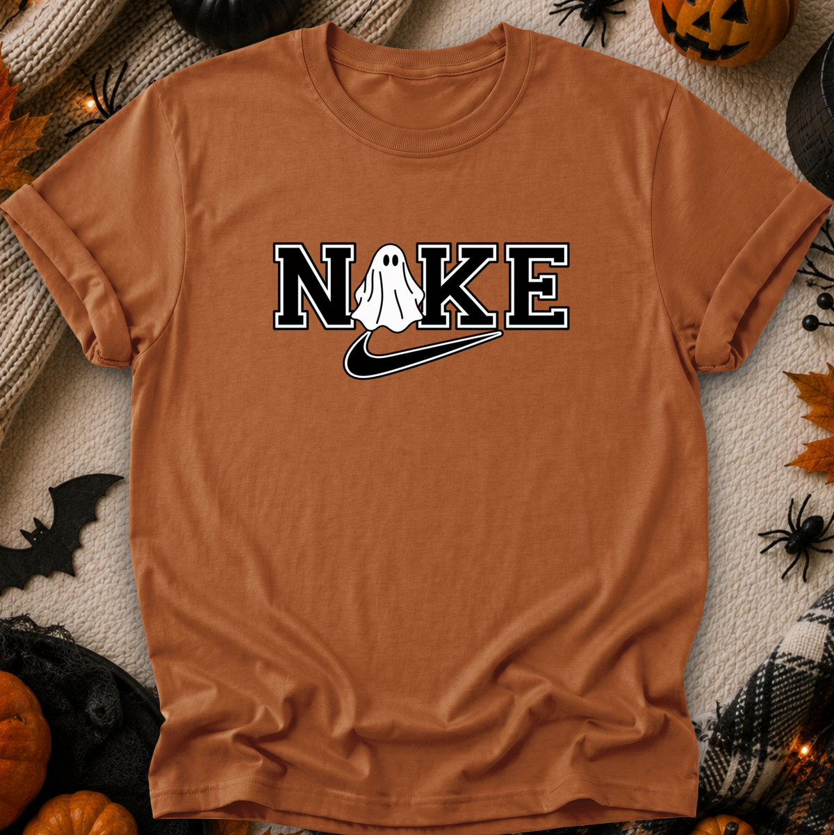 A rust-orange short-sleeve t-shirt laid flat with Halloween-themed props around it. The shirt has a graphic reading 'NIKE' with a white ghost and a black swoosh replacing the 'I', styled like the Nike logo.