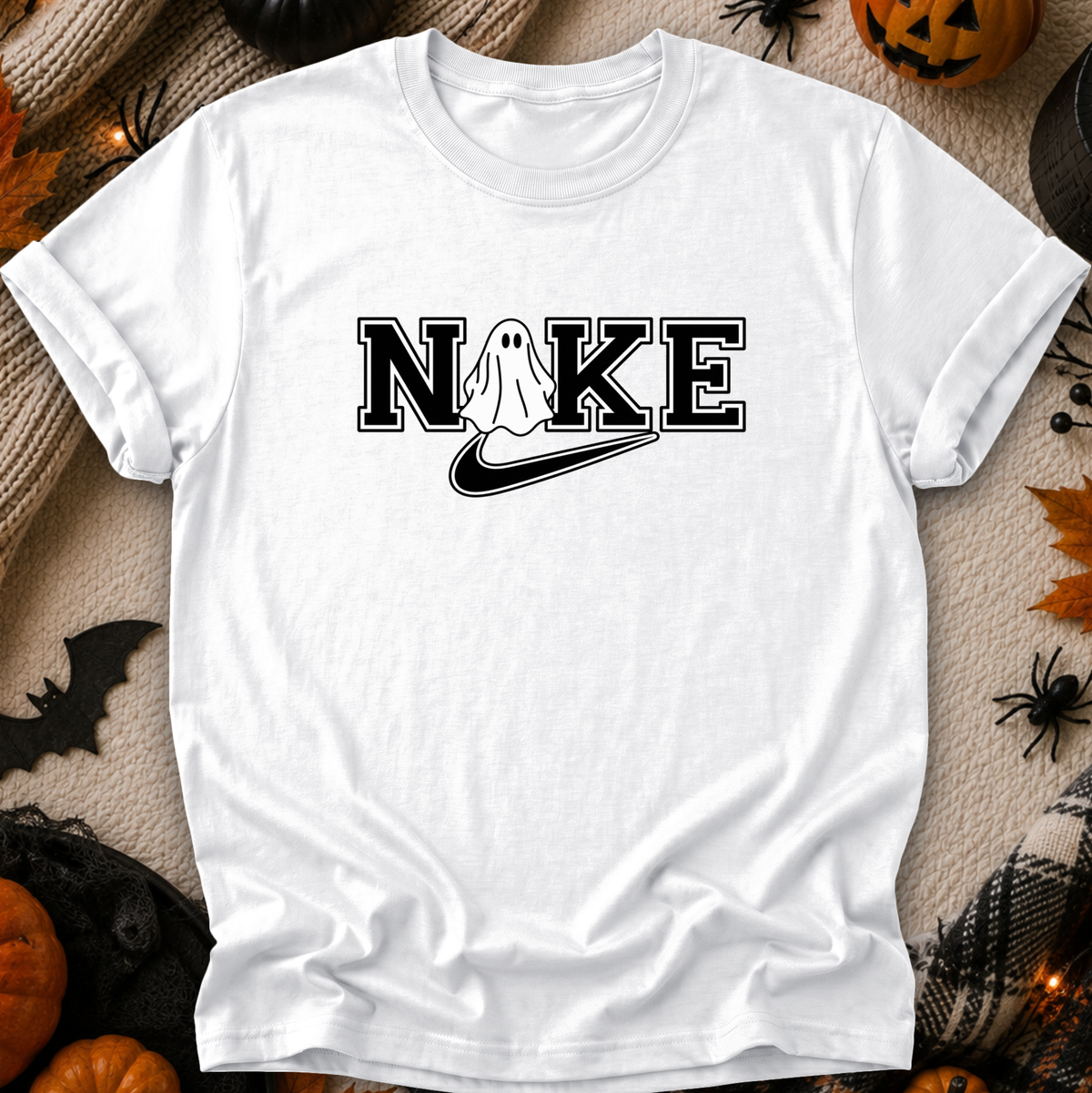 A white short-sleeve t-shirt with a black graphic print reading 'NIKE' where the 'I' is replaced by a ghost and a swoosh, styled in a flat-lay with Halloween decorations.