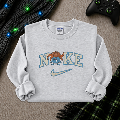 A light heather gray sweatshirt featuring a 'NIKE' graphic with a cartoon pokemon gloomy and a Nike swoosh, styled flat-lay on a textured beige surface with a black controller, a knit sweater, and a plaid scarf as props.