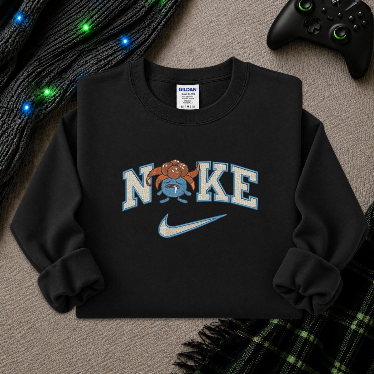 A black crewneck sweatshirt laid flat on a textured beige surface, featuring a graphic that reads 'NIKE' with a cartoon pokemon gloomy wearing a Nike swoosh, styled with a black controller, knitted cable lights, and a plaid blanket.