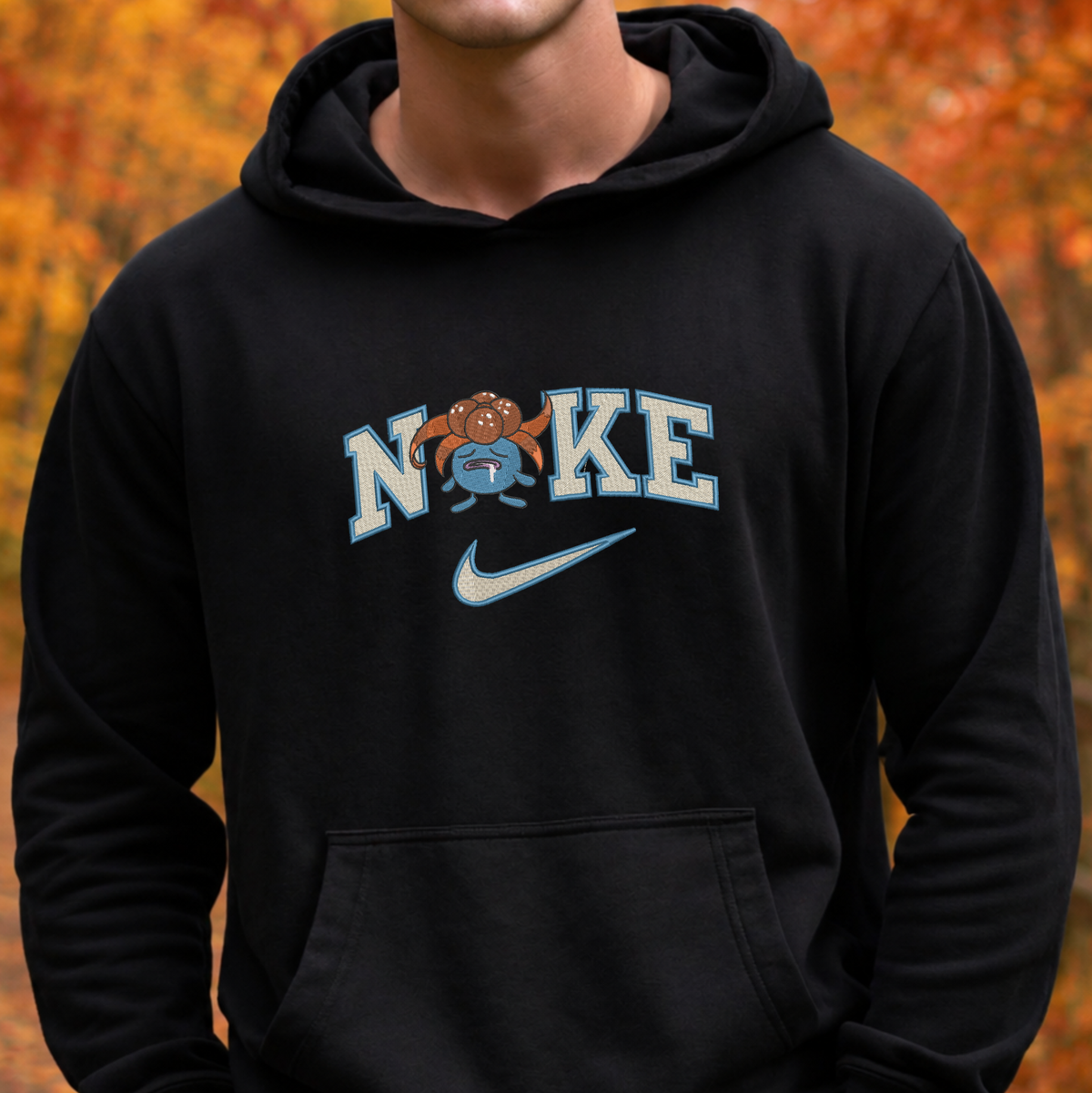 A person wearing a black pullover hoodie with a front kangaroo pocket and a large 'NIKE' graphic across the chest. The graphic features light blue varsity-style lettering, a small cartoon mascot, and a light blue Nike swoosh.