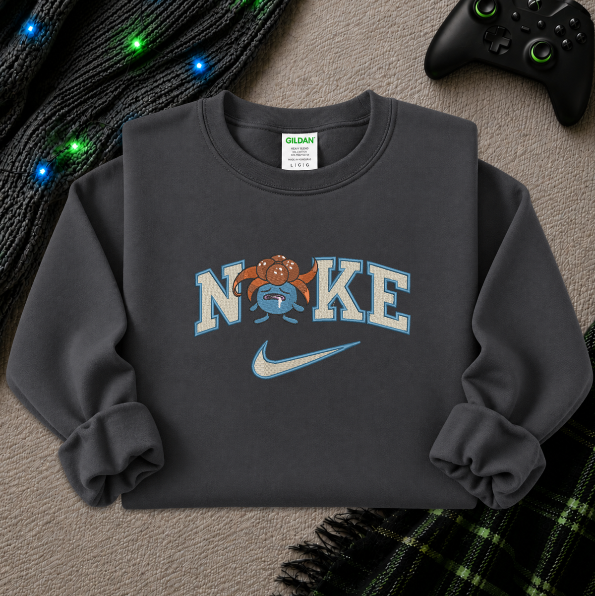 A dark gray crewneck sweatshirt laid flat on a textured beige surface, featuring a custom graphic that reads 'NIKE' with a cartoon pokemon gloomy and a Nike swoosh. A Gildan brand tag is visible inside the collar.