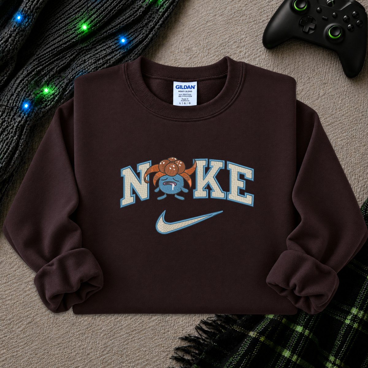 A dark brown crewneck sweatshirt featuring a 'NIKE' graphic with a cartoon mascot and a Nike swoosh, styled flat-lay on a textured beige surface with a game controller, a black knit blanket with colorful lights, and a plaid blanket.
