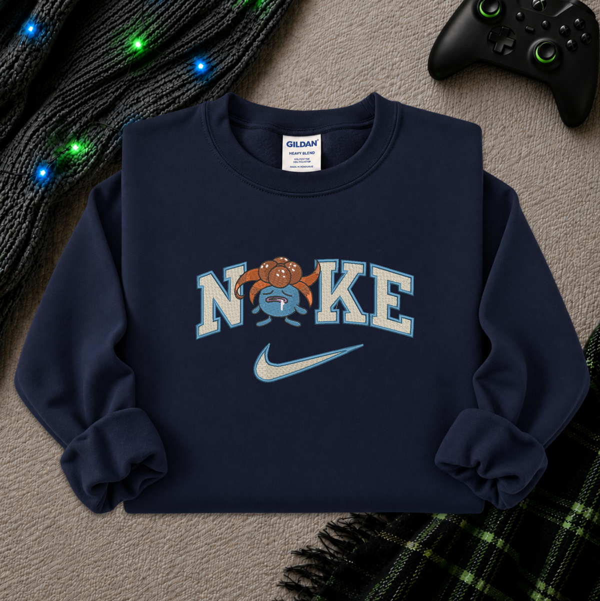 A navy blue crewneck sweatshirt with a graphic print reading 'NIKE' featuring a cartoon pokemon gloomy and a Nike swoosh, styled flat-lay on a textured gray surface with a black controller, knit sweater, and plaid blanket nearby.