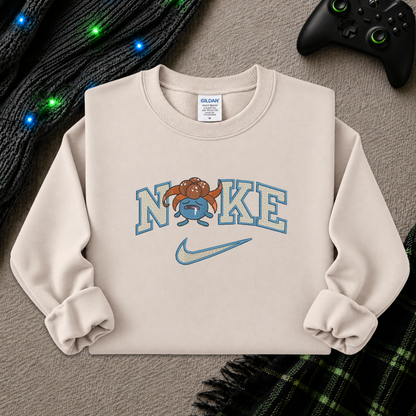 A cream-colored crewneck sweatshirt featuring a 'NIKE' graphic with a cartoon gloomy and a light blue Nike swoosh, styled flat-lay on a gray textured background with a black controller, a knit sweater, and holiday lights.