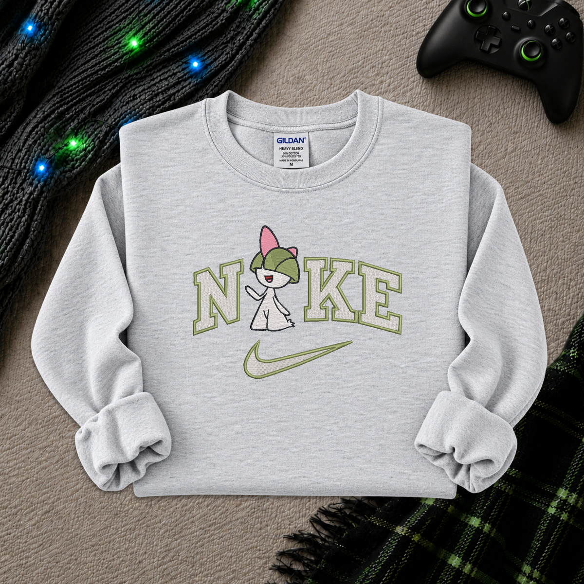 A light gray sweatshirt featuring a graphic of a pokemon Raltz cartoon character with a green hat, standing in front of the word 'NIKE' in olive green letters, with a green Nike swoosh underneath. The sweatshirt is laid flat on a textured beige surface with a black controller, a black knit sweater with blue fairy lights, and a green plaid blanket as props.