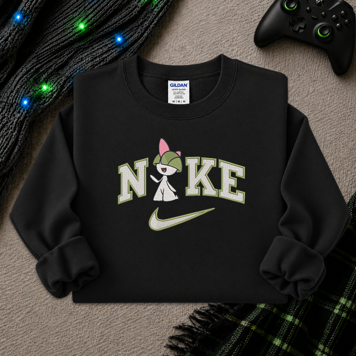 A black crewneck sweatshirt featuring a graphic print of the word 'NIKE' with a pokemon Raltz cartoon character inside the letter 'I', a green Nike swoosh, and a Gildan brand tag visible inside the collar. The sweatshirt is styled in a flat-lay arrangement on a textured gray surface with a black cable, a game controller, and a green plaid blanket.