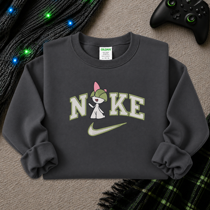A dark gray crewneck sweatshirt featuring a pokemon Raltz cartoon character in a green helmet and pink hair bow replacing the 'O' in 'Nike', with 'NIKE' printed in white and green varsity letters and a green swoosh. The sweatshirt is styled flat on a textured beige surface with a black game controller, a black knit cable with blue lights, and a green plaid blanket.