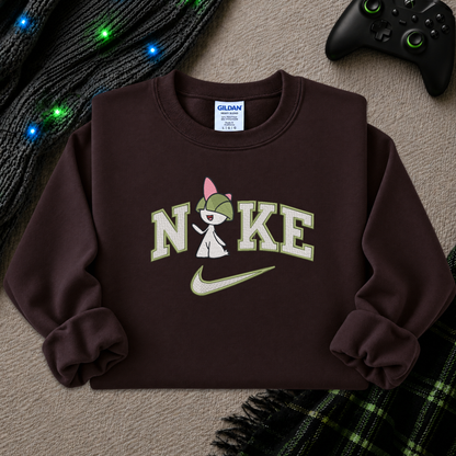 A dark brown crewneck sweatshirt featuring a custom graphic print of the word 'NIKE' with a cartoon character inside the 'I', a light green Nike swoosh, and a visible Gildan brand tag inside the collar. The sweatshirt is styled in a cozy flat-lay setting with a black knit sweater, glowing blue fairy lights, a black video game controller, and a green plaid blanket on a textured beige carpet.