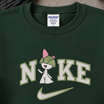 A close-up of a dark green crewneck sweatshirt featuring a custom graphic. The graphic displays the word 'NIKE' in white varsity-style letters with a light green outline, where the 'I' is replaced by a pokemon raltz cartoon character wearing a green cap.