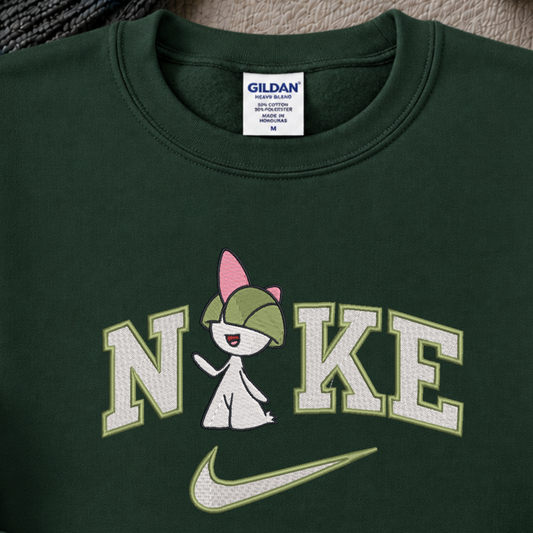 A close-up of a dark green crewneck sweatshirt featuring a custom graphic. The graphic displays the word 'NIKE' in white varsity-style letters with a light green outline, where the 'I' is replaced by a pokemon raltz cartoon character wearing a green cap.