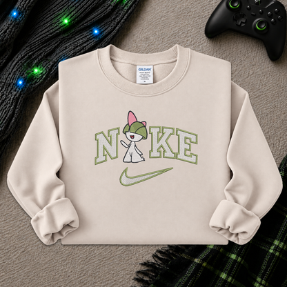 A beige crewneck sweatshirt featuring a graphic of a pokemon Raltz cartoon character wearing a green cap and holding a sign that says 'NO KE' with a green Nike swoosh underneath. The sweatshirt is styled flat on a gray textured surface with a black knit sweater, colorful fairy lights, and a black game controller in the background.