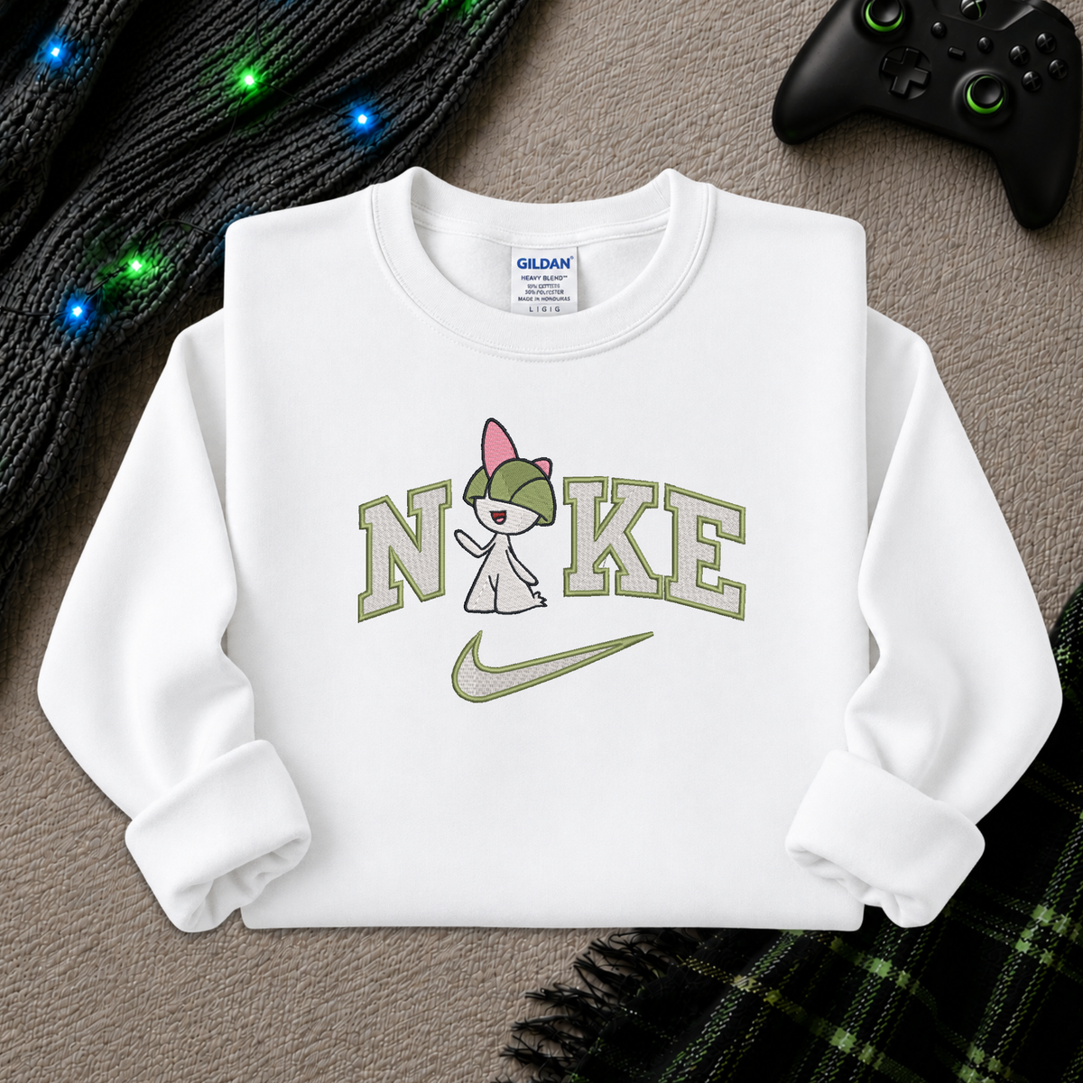 A white long-sleeve crewneck sweatshirt laid flat on a textured gray surface, featuring a graphic print of the word 'NIKE' styled with a pokemon Raltz cartoon character wearing a pink beanie and a green swoosh. The neck label shows Gildan branding.