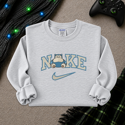 A light gray crewneck sweatshirt featuring a 'NI KE' graphic with a cartoon pokemon snorlax character and a Nike swoosh, styled flat-lay on a textured beige surface with a game controller, knitted sweater, and plaid blanket.