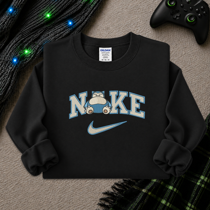 A black crewneck sweatshirt laid flat on a textured gray surface, featuring a graphic that reads 'NIKE' with a cartoon pokemon snorlax inside the letter I and a light blue Nike swoosh underneath. A Gildan brand tag is visible inside the collar.