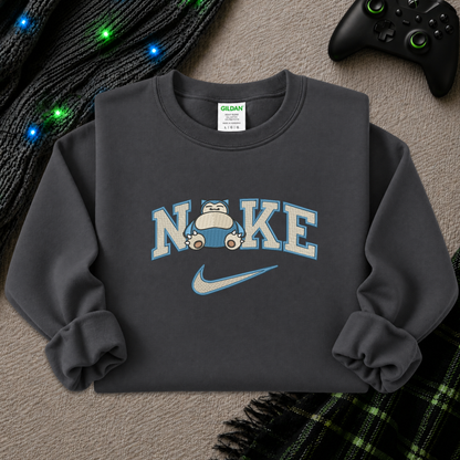 A dark gray crewneck sweatshirt featuring a 'NI KE' graphic with a cartoon pokemon snorlax character and a Nike swoosh, styled flat-lay on a textured beige surface with a game controller, knitted sweater, and plaid blanket.