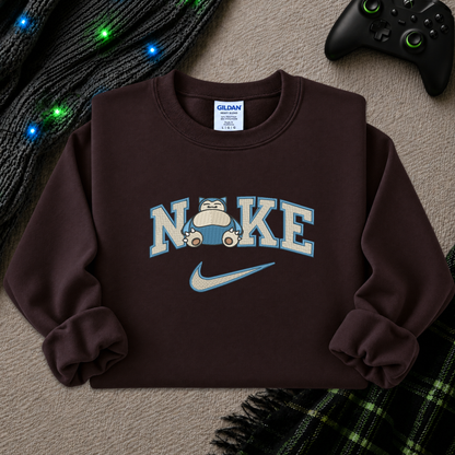 A dark brown crewneck sweatshirt featuring a 'NI KE' graphic with a cartoon pokemon snorlax character and a Nike swoosh, styled flat-lay on a textured beige surface with a game controller, knitted sweater, and plaid blanket.