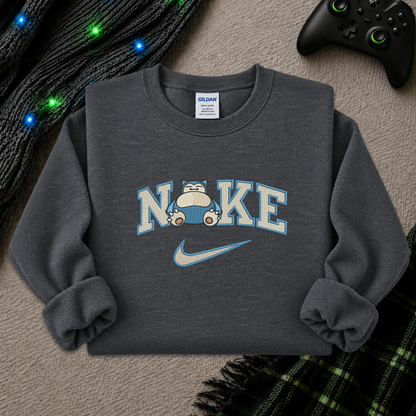 A dark gray crewneck sweatshirt featuring a 'NI KE' graphic with a cartoon pokemon snorlax character and a Nike swoosh, styled flat-lay on a textured beige surface with a game controller, knitted sweater, and plaid blanket.