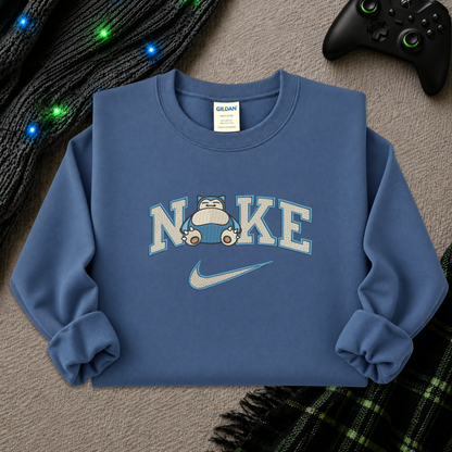 A blue crewneck sweatshirt featuring a 'NI KE' graphic with a cartoon pokemon snorlax character and a Nike swoosh, styled flat-lay on a textured beige surface with a game controller, knitted sweater, and plaid blanket.