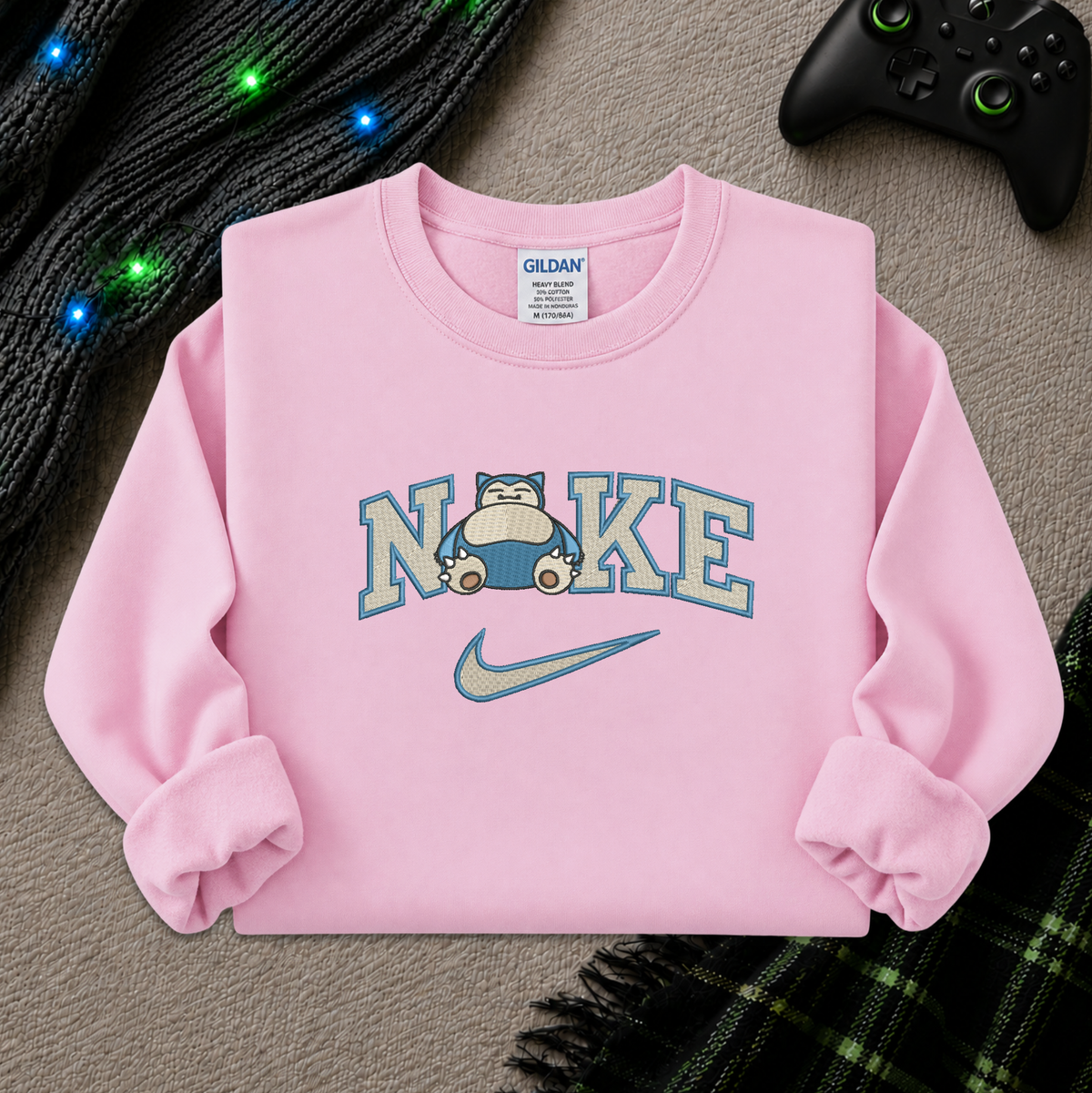 A light pink crewneck sweatshirt featuring a 'NI KE' graphic with a cartoon pokemon snorlax character and a Nike swoosh, styled flat-lay on a textured beige surface with a game controller, knitted sweater, and plaid blanket.